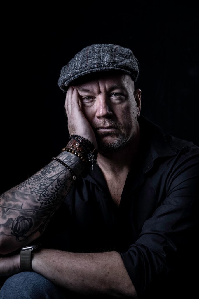 Jack Cartwright resting his face on his hand, in a flat cap and black shirt